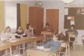 En skolklass sitter i sina bänkar i ett klassrum, Brattåsskolan 1970- till 80-tal. Närmast till vänster ses genom fönstren att landskapet är vintervitt av snö. I bakgrunden till vänster ses ett högskåp med en jordglob placerad överst. Till höger ses 