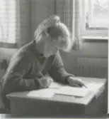 En elev sitter vid en skolbänk och skriver, klassrum i Brattåsskolan 1960-tal. Flickan har håret uppsatt i hästsvans.