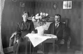 Par på soffa bakom blomsterprytt bord med broderad duk, 1920-tal