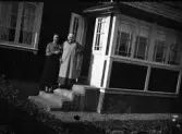 Två kvinnor står på trappan utanför ett hus med glasveranda. Kanske avser anteckningen 
