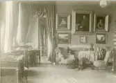 Foto visar ett rum med bl.a soffa, fåtöljer, bord, stora målningar med breda ramar, draperi mm. inifrån Gysinge herrgård.