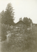 Liten stuga/uthus med gärdsgård och vedtrave omgiven av skog, troligen i trakterna runt Gysinge.