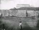 Reprokopia. Okänd dam framför fästningen sedd från söder. Notera sorgbandet på hennes vänstra arm. Av klädedräkten att döma är bilden från 1910-talet. På södra fästningsmurarna ser det ut som någon typ av byggnadsställningar kommit upp. Renorveringen av fästningen drevs som s k AK-arbete mellan åren 1922-1924. Möjligen är bilden från denna tid.