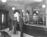 Interiörbild från expeditionslokalen i Varbergs sparbank. Mannen bakom disk till höger heter Arvid Adolfsson. Fönstren i bakgrunden vetter mot öster. Till en början hade man banklokaler på bottenvåningens östra del medan Hallandsbanken och Göteborgsbanken hyrde lokaler i den västra.