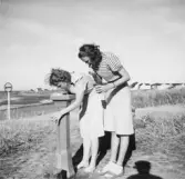 Två kvinnor som borstar tänderna vid en drickfontän i strandmiljö. I bakgrunden ses badstugor. Troligen badgäster vid Stora Apelvikens camping.