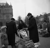 En torgmadam som säljer pepparrot och virar in dem i tidningspapper, antagligen från Fjärås. I förgrunden en liten pojke med öronlapparna nedfällda på kepsen och i bakgrunden en blomsterförsäljare. Bilden är tagen efter 1937 men före 1944.