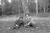 Två herrar i kostym, överrock och hatt sitter vid ett träd i skogen, 1920-tal