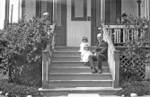 Man med två små barn sitter i trappan medan fler personer vistas på verandan, 1920-tal