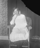 Konfirmand i vit klänning med vita handskar sittande i korgstol i hemmet, 1920-tal