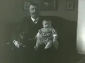 Handlanden Olof Larsson sitter i en soffa tillsammans med en hund och ett barn, troligen sonen Erik Sören f. 1909.