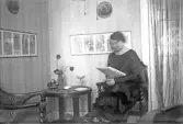 Kvinna med pälskantad klänning sitter i läshörnan och läser en tidning, 1920-tal