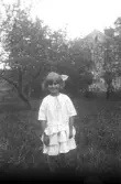 Porträtt av liten flicka i vit klänning och hårrosett i trädgård, 1920-tal