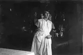 Kvinna i klänning och förkläde i mörkt rum, 1920-tal