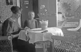 Kvinna i korgstol broderar med en pojke bredvid sig, 1920-tal