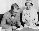 Herr Malmrén skriver under första gireringskortet från kontot
(överföring till girosparkonto), fru Inga ser på. Lokal:
postsparbankschefens tjänsterum.