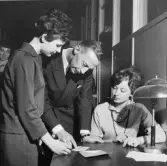 För att underlätta för utbetalningskassorna, i Postbankens
korridorer, hade man satt in speciella legitimationskontroller - här
Ilja Forsblom t.h. Systemuppfinnaren byråsekreterare Yngve Cassius
hjälper fru Gunvor Boman på bankbokföringen tillrätta.