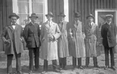 Sju herrar i hatt och ytterrock står i rad framför trähus, 1920-tal