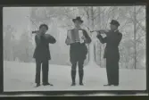 Albin, Gunnar och Johan Karlsson, Gimmersta, står utomhus i snön och spelar musik. En på dragspel och de andra två på fiol.