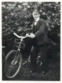 Fjärdingsman Karl Gustaf Johannesson i Aneboda, ca 1925-30, poserar med en cykel i en trädgård.
Något suddigt foto.