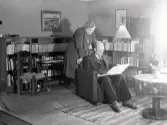 Museichef Albert Sandklef med frun Karin Sandklef i bostadens bokhörna. Albert läser och Karin står bakom honom. Fotografiet är taget i Artillerikasernen på Fästningen.