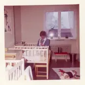 Fotografi på en barnsköterska som matar en baby med nappflaskan i barnhemmets sovrum, påsken 1962, på Lilla Källbäck.