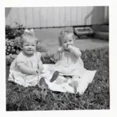 Fotografi på två små flickor med klänningar som sitter på en filt i gräset, troligen vid Lilla Källbäck.