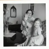 Fotografi på en mamma, ett spädbarn och ett litet större barn som sitter i en soffa i ett vardagsrum.