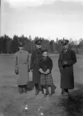 Tre uniformerade herrar (varav en polis ?) och en flicka med skolmössa står på en äng. I bakgrunden skymtar en militärbarack (?).