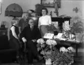 Foto av en kostymklädd man som sitter i ett vardagsrum, omgiven av sin familj och mängder av blommor.
50-årsdag (?).