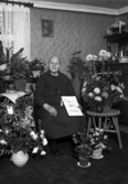 Foto av en kvinna i klänning som sitter i ett vardagsrum, omgiven av blommor. 
Trol. fru Anna Kristina Kristoffersson, f. Karlsson (12 december 1861-1947), Villansdal, Hov, Växjö.
Källa: Sveriges Dödbok 1901-2009.