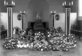 Interiörfoto från (trol. kapellet på Tegnérkyrkogården) inför en begravning. Man ser en mörk kista omgiven av höga kandelabrar och blommor.