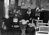Foto av en kvinna i klänning som sitter bredvid en radio i ett vardagsrum, omgiven av blommor.