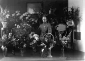 En man i uniform sitter i ett vardagsrum, omgiven av mängder av blommor.
