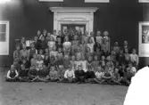 OBS! Skadat i nedre högre hörnet.
Foto av en skolklass med lärare, som samlats utanför skolan.