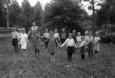 Foto av en skolklass (?) med lärarinna, som dansar ringdans i en trädgård.