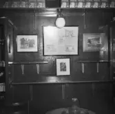 Interiör sannolikt av kontoret i Oscar Pehrsons speceriaffär. Väggparti med inramade fotografier av butiken över väggfasta hyllor mellan glasskåp. På hyllan upptill står paket med 