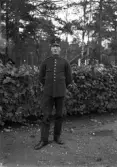 Foto av en man i mörk militäruniform, stående vid en häck.