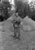 Foto av en ung man i uniform framför några kamouflagemålade tält.
I 11, Växjö