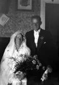 Foto av ett brudpar i ett vardagsrum. Hon är klädd i vit brudklänning med slöja och pärldiadem. Han bär frack.