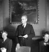 Sammanträde på Stadshotellet, 1943. 
En äldre man håller tal, stående mellan två sittande damer.