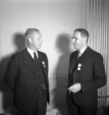 Sammanträde på Stadshotellet, 1943. 
Två herrar i kostym och med medaljer på kavajslaget pratar med varandra.