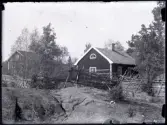 Vy över två byggnader i Stensjö by. I förgrunden en karg berghäll över vilken det löper en gärdesgård byggd med vidjor. I mitten av bilden syns en grind. Till höger om grinden syns en stege på gärdesgården. Byggnaden innanför är Lillstugan, vilken har ett lunettfönster på gaveln. Bakom en ridå av unga träd syns Stensiögården.
