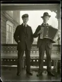 Eugén och Bengt Stensiö står på en veranda. Båda bär mörk kostym med slips och blanka svarta skor. Eugén bär studentmössa och Bengt en hatt och glasögon. Han håller i ett dragspel. 
På verandan står en ljust målad soffa i trä. Verandans snickarglädje syns bakom soffan samt till höger i bild. Bostadshuset har ett smyckat listverk.