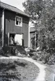 Morfars hus i Stensjö by. En trappa leder upp till den öppna dörren. Vid takdroppet syns en hängränna i trä. I dörröppningen står Judit Karlsson och framför henne sitter Martin Karlsson. Judit bär en kortärmad klänning eller blus med ett bredaxlat smutsigt förkläde med påsydda band eller broderi. Martin bär en vit skjorta med mörk väst och mörka byxor.  Till höger i bild syns fruktträd och en gärdesgård bunden med vidjor. På den hänger ett tygstycke. I förgrunden syns en upptrampad stig i gräsmattan. På trappan står en hink och till höger om trappan en större balja samt en hink, alla i zink.