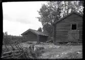 Vy över bygata och två uthus i Stensjö by. En mindre byggnad med en liten lucka vid högra gaveln och en större byggnad i rundtimmer med en liten lucka mitt på byggnaden. Marken framför byggnaderna är upptrampad och visar körspår. I kanten av vägen syns en gammal huggen stubbe. Till vänster syns två gärdesgårdar i trä bundna med vidjor. Bakom byggnaderna syns kronan av en stor asp med möjliga spår av hamling.