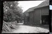 Två barn utanför Magasinet i Stensjö by. Till högerär en flicka med mörk klänning och vitt smutsigt förkläde. Hon har vara bara ben och skor. På en kärra parkerad i byggnaden sitter en pojke. han bär en uppknäppt mörkare skjorta över en ljusare skjorta, mörka byxor och skor. Utanför byggnaden löper en väg. Till höger i bild syns en hög med klena träd eller grenar. Längs vägen löper ett staket med fruktträd innanför.