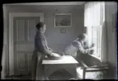 Tilia Andersson står vid ett bord med händerna i ett såll och rensar något. Hon bär en mörk rutig klänning, randigt förkläde och en ärmlös kofta med knappar och virkad spets i kanterna. Hon har håret i en knut i nacken. Lutad vid fönstret syns Anna Karlsson i rutig klänning och ljust randigt förkläde. Hon håller ett föremål med snören i händerna. Fönstret är inramat med tunna ljust randiga gardiner. I kökssoffan ligger en duk eller papper fylld med samma sak som rensas i sållet i Tilias händer, samt en pappesrulle och ett tefat med snöre.
Bakom Kvinnorna syns en spegeldörr målad i olika färger. På väggarna sitter en blommig tapet samt två tavlor. Den större har ett stadsmotiv och den mindre under är möjligen ett porträttfotografi. På väggen längst till höger syns en almanacka med datumet 8, möjligen augusti.