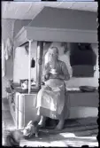 Pigan Tilda Karlsson sitter barfota vid spisen med en kopp kaffe på fat med tesked i. Hon har grånande hår i knut, en blus med hög krage, smalrandig kjol och ett smutsigt ljusrandigt förkläde. På spisen och bänken bakom henne syns kaffekannor och olika typer av kärl. Till höger om henne hänger ett par yllestrumpor och till vänster grytlappar. Till höger syns en ugnslucka till en brödugn. Gjutjärnsspisen till vänster har en tank för uppvärmt vatten. På trasmattorna framför henne på golvet sitter två katter, den ena lapar ur en skål.