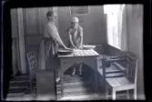 Tilia Andersson och Anna Karlsson står vid ett bord med och bakar kakor. Hon bär en ljus randig klänning, randigt förkläde. Hon har håret i en knut i nacken. Anna har en kortärmad klänning med ett ärmat förkläde med fickor över. Hon bär skor med slejf. 
Fönstret är inramat med tunna ljust randiga gardiner. I kökssoffan står ett krus och längre bort ligger en tidning. 
På bordet står två bakkärl och handduk med ogräddade kakor. På golvet ligger trasmattor och mot en stol står en plåt lutad.
Bakom kvinnorna syns en spegeldörr målad i olika färger. På väggarna sitter en blommig tapet samt två tavlor. Den större har ett stadsmotiv och den mindre under är möjligen ett porträttfotografi. På väggen längst till höger syns en almanacka med datumet 15 måndag angivet.