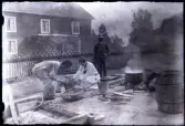 Två män och en kvinna bland en mängd kärl och redskap i Stensjö bykärna. Från höger Eugén Stensiö och Aina Stensiö. I förgrunden ligger en vedbock med en såg. Eugén häller något i hinken som Aina rör i. Framför står en flätad korg. I bakgrunden står en okänd man. Till höger om honom syns en rykande kittel och en stor tunna.
I bakgrunden syns ett staket med vita spetsar samt Stensiögården. Byggnaden har en veranda överväxt med klätterväxter. Bostadshuset bakom staketet har en veranda med en klättrande perenn. Byggnaden har stående locklistpanel på övervåningen och liggande panel på nedervåningen.